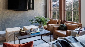 Ein modernes Wohnzimmer mit großem Flachbildfernseher an einer strukturierten Wand, Ledersofas, einem Couchtisch mit Büchern und einer Vase, und Blick auf eine Berglandschaft durch große Fenster.