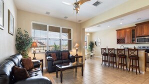 A spacious living room with a dark leather sofa, a coffee table, and a ceiling fan. Adjacent is a kitchen with wooden cabinets and a bar area with stools.