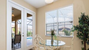A dining area with a glass table and white metal chairs, a potted plant, and a view through a window with blinds.