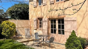 Una casa de dos pisos con un patio amoblado, que incluye una mesa y sillas, rodeada por un césped cuidado y un pequeño árbol.