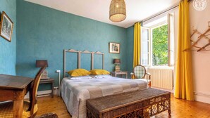 Une chambre à coucher comprenant un lit, des tables de chevet, une chaise, un banc en osier et une fenêtre avec des rideaux.
