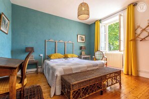 Une chambre à coucher comprenant un lit, des tables de chevet, une chaise, un banc en osier et une fenêtre avec des rideaux.