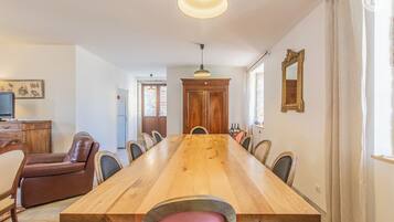 A spacious dining area with a long wooden table, surrounded by chairs, a brown leather sofa, and a television on a wooden cabinet.