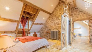 A bedroom with a sloped ceiling, stone walls, a bed, a radiator, and a window with curtains.