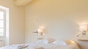 Una cama bien tendida con sábanas y toallas blancas, suelo de madera, puerta blanca e iluminación montada en la pared.