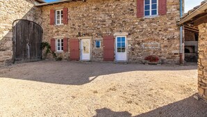 Un edificio de piedra con un patio de grava, una puerta de madera grande y contraventanas rojas en las ventanas.