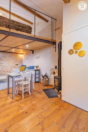 Een gezellige kamer met houten vloer, een stenen muur, een eettafel met stoelen, een bank, een open haard en een vide met houten plafond.