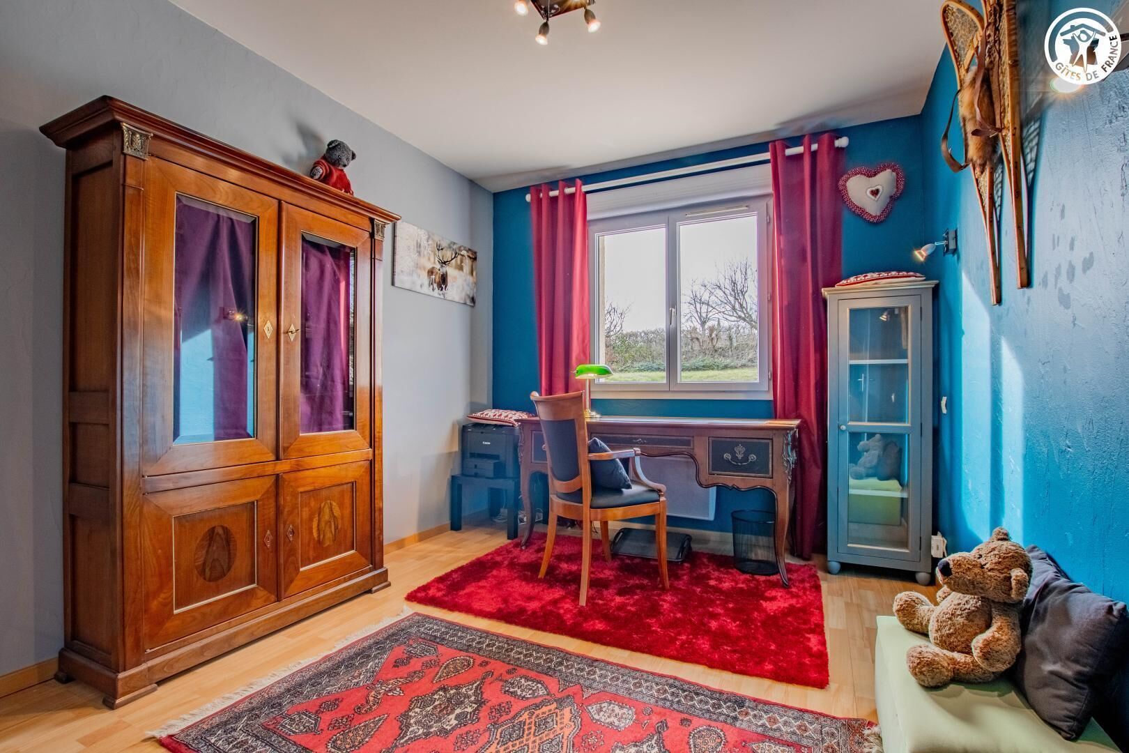 A room with a wooden wardrobe, a desk with a chair, a red rug, a window with curtains, and a teddy bear on a sofa.
