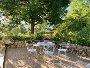 A patio with a round table and white metal chairs, surrounded by a stone wall and lush greenery.