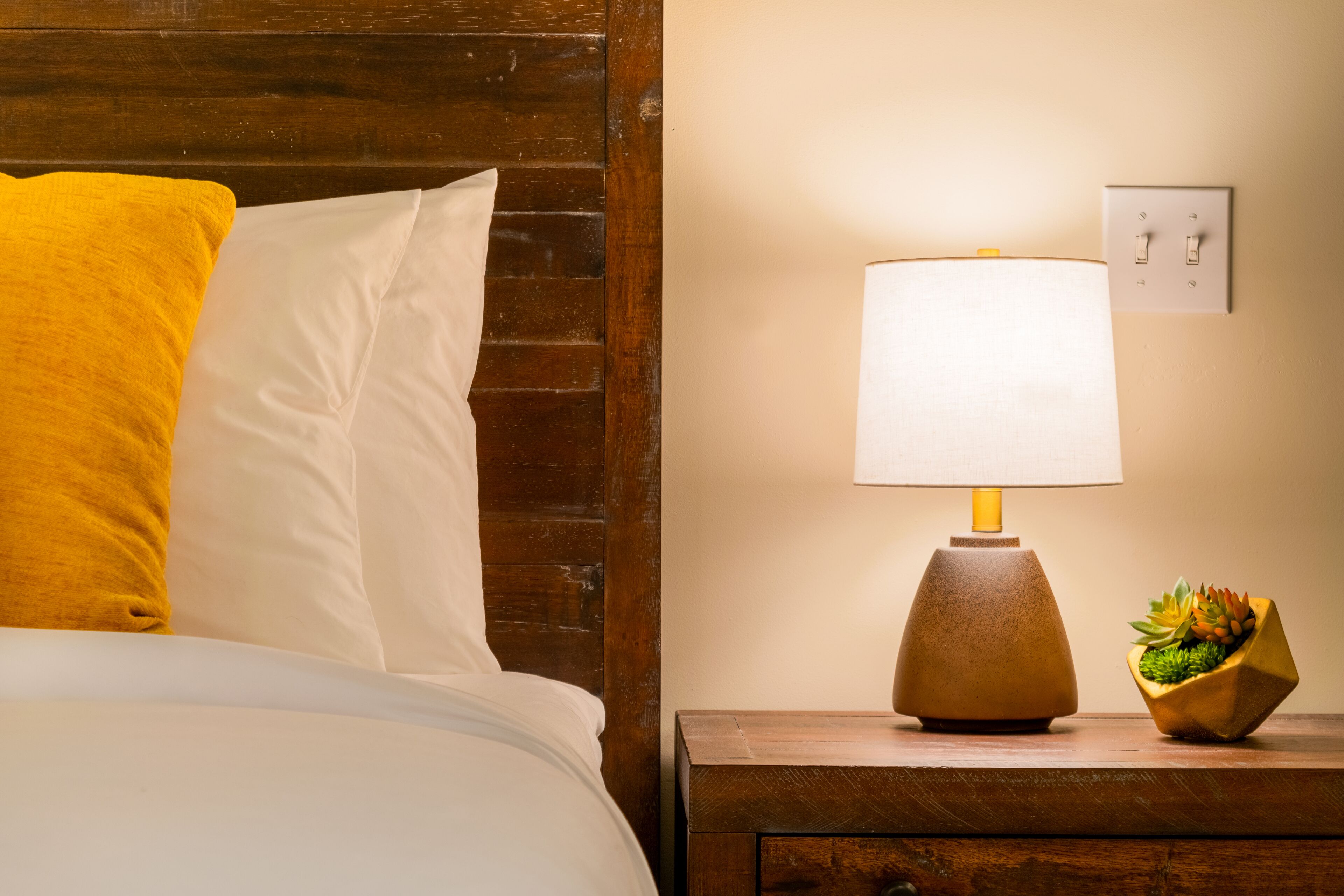 A neatly made bed with white and yellow pillows, a wooden nightstand with a lamp, and a small plant.