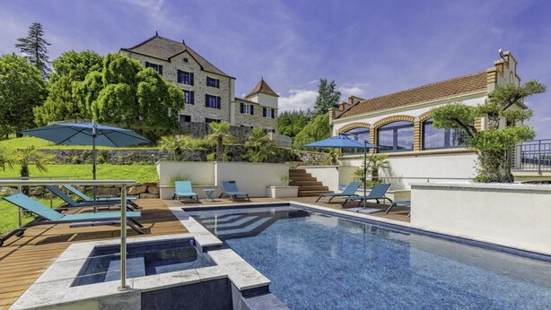 A swimming pool with lounge chairs and umbrellas, surrounded by a stone building and greenery.