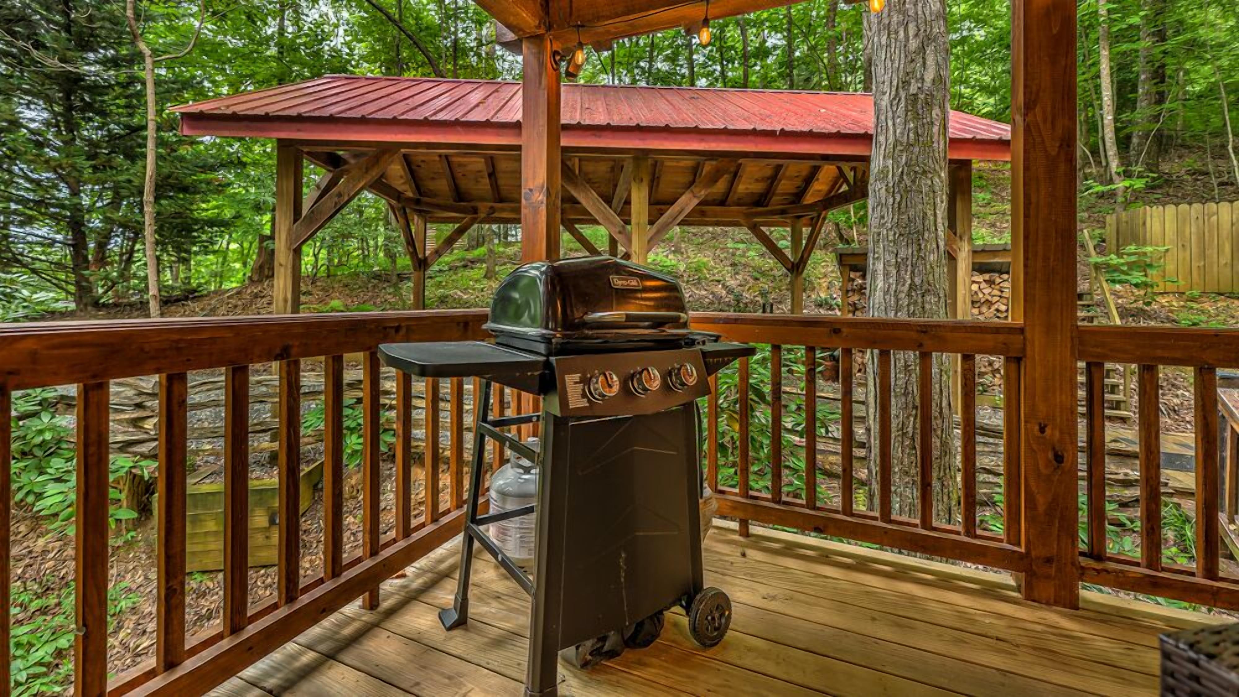 A covered wooden deck with a barbecue grill and a table, surrounded by a wooden railing and trees.