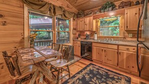L’intérieur d’un chalet en bois comprend une table à manger, des chaises, une cuisine avec des meubles en bois, une fenêtre donnant sur la verdure et un tapis sur le sol.