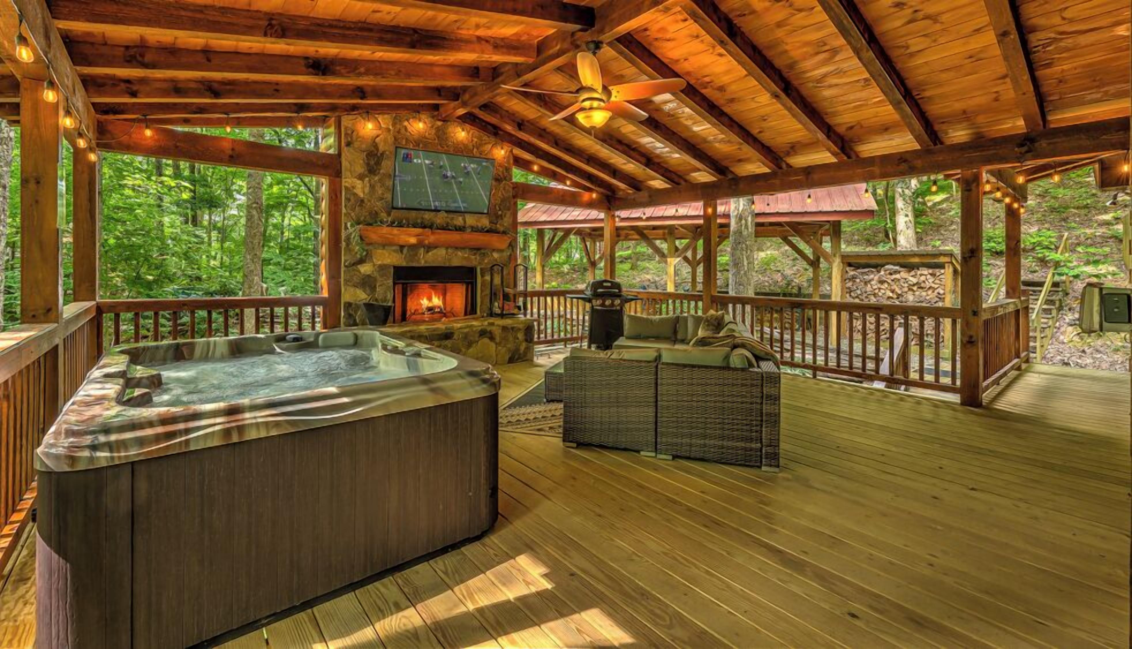 A wooden deck with a hot tub, outdoor seating, a stone fireplace, and a TV mounted on the wall.