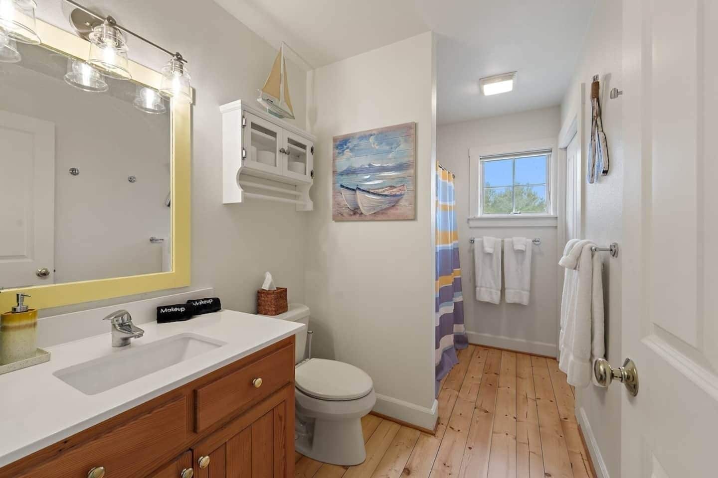 Une salle de bain avec un plancher en bois, un lavabo blanc, un miroir et un tableau représentant un voilier.
