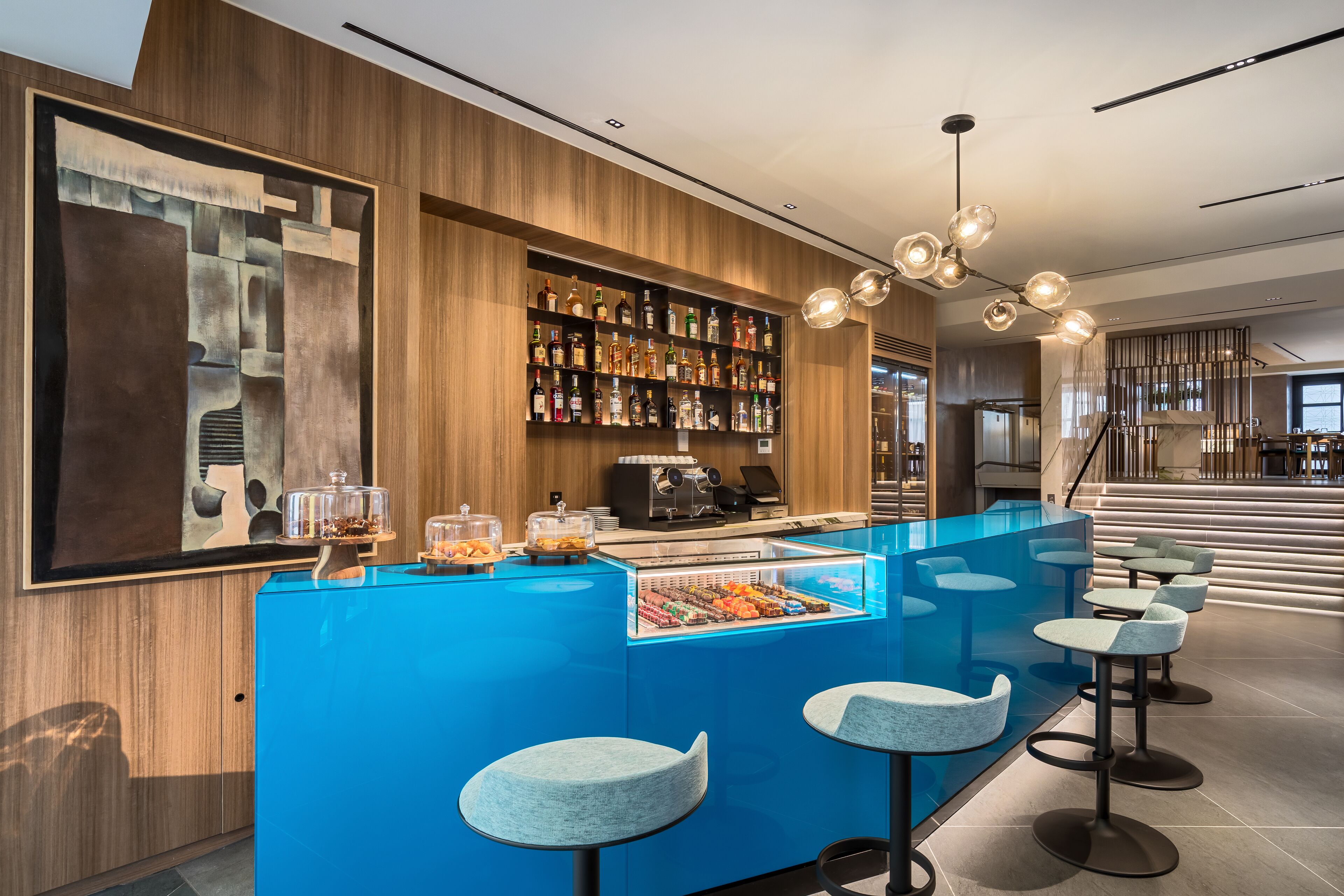 A modern bar area with a blue counter, bar stools, a coffee machine, and a variety of liquor bottles.
