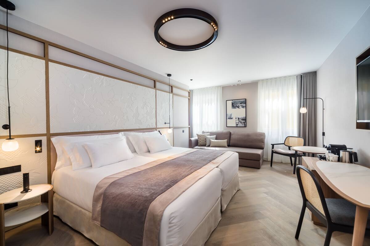 A modern hotel room with a large bed, a sofa, a dining table, and a desk.