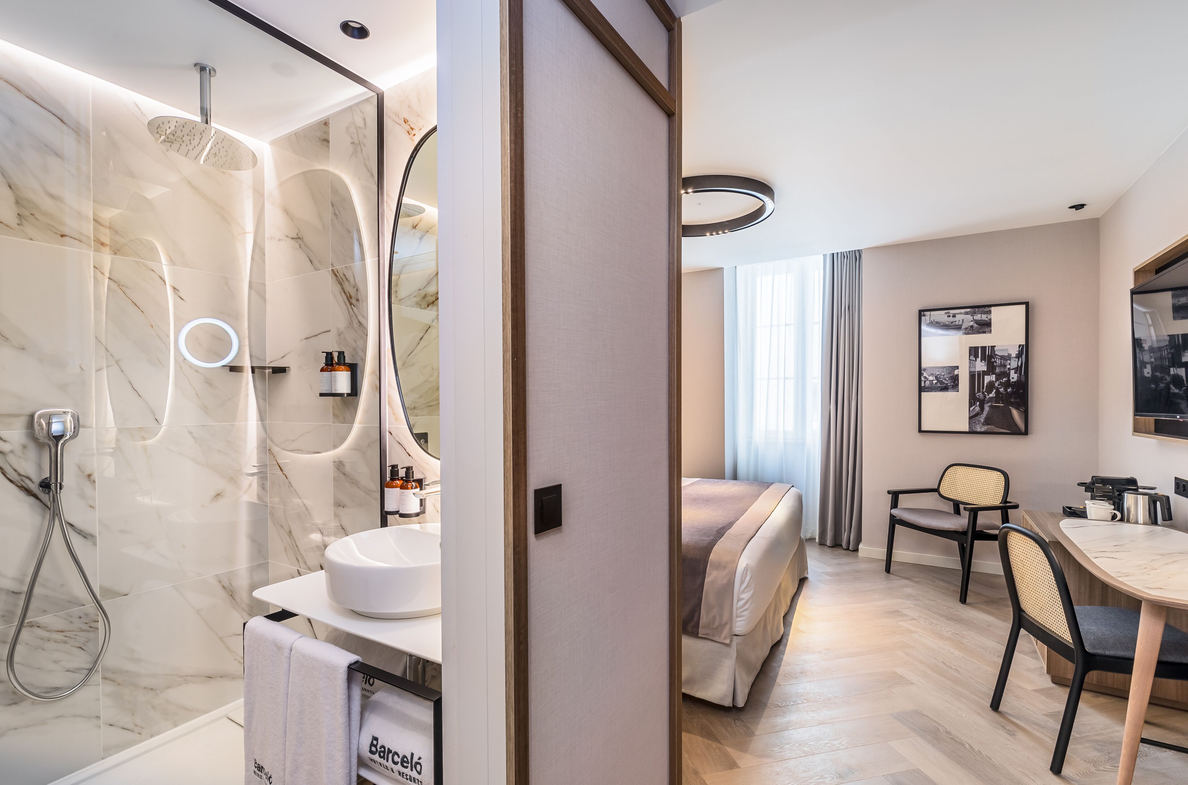 A modern bathroom with a marble shower, a sink, and a toilet. There is a bed with a headboard and a bedside table with a lamp. The room has a wooden floor and a large window with curtains.
