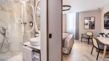 Un baño moderno con ducha de mármol, un lavabo y un inodoro. Hay una cama con cabecera y una mesita de noche con lámpara. La habitación tiene suelo de madera y un ventanal con cortinas.