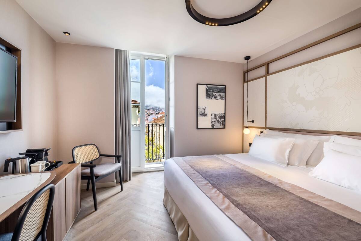 A hotel room with a bed, a desk with a chair, a television, and a balcony with a view of the city.