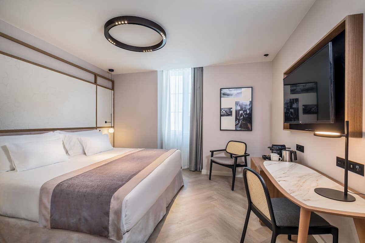 A hotel room with a bed, a desk, a chair, a TV, and a window with curtains.