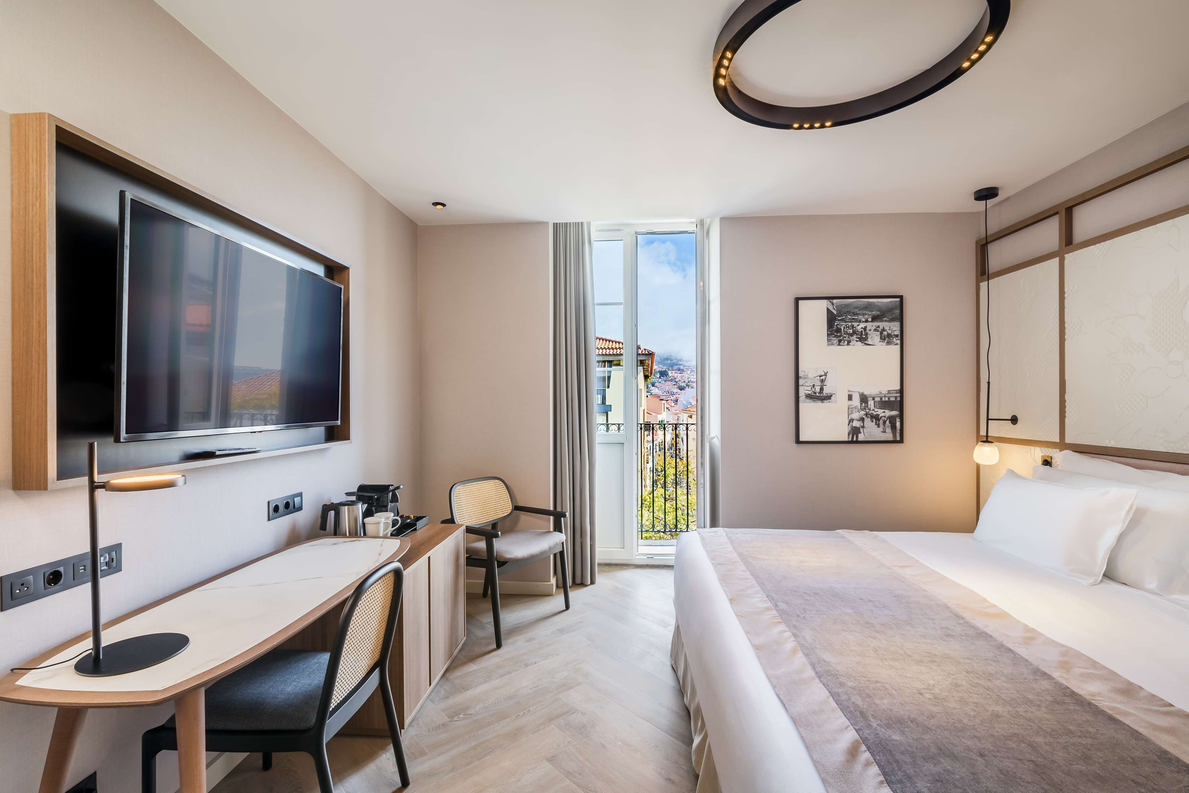 A modern hotel room with a large bed, a desk, a flat-screen TV, and a balcony with a city view.