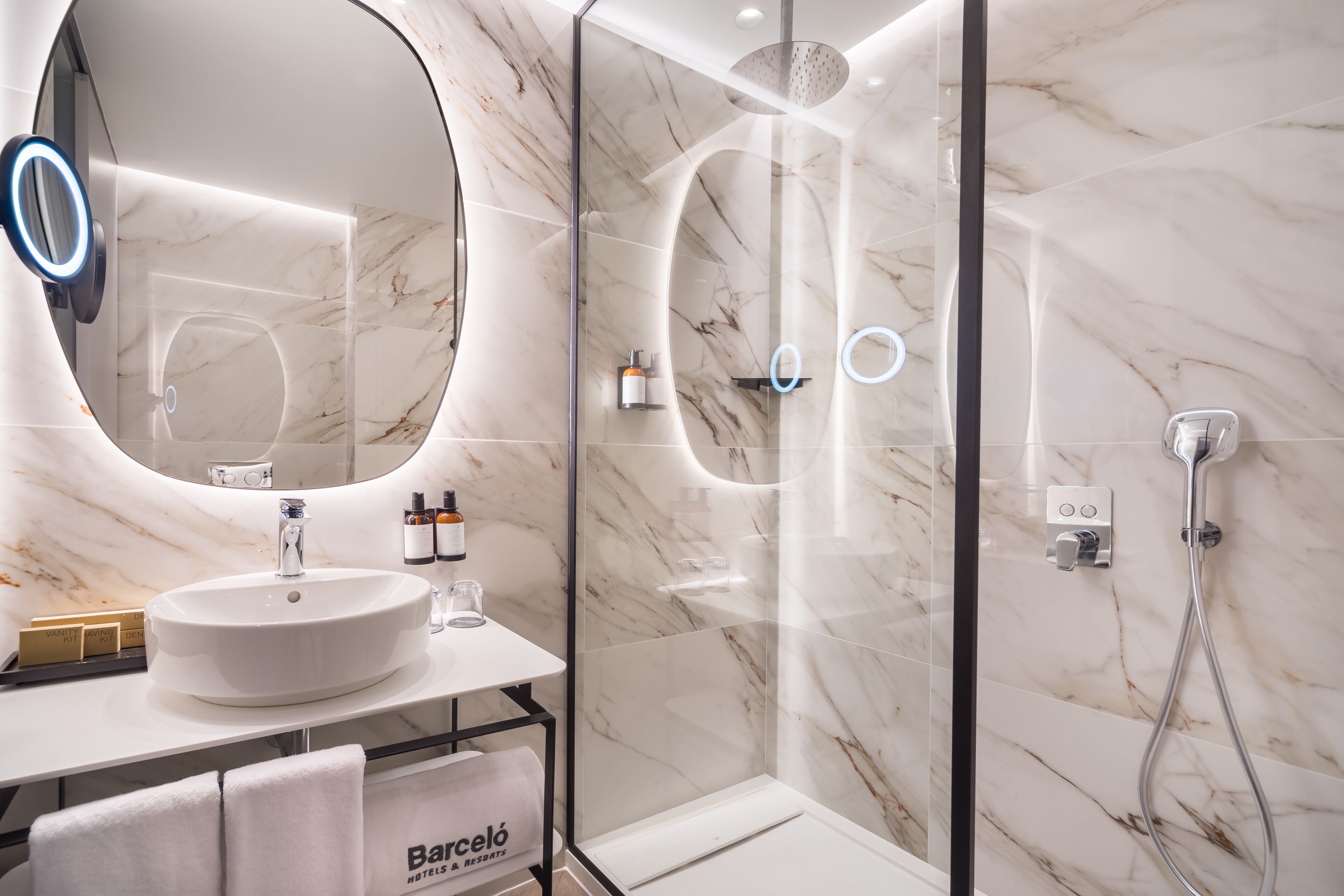 Un baño moderno con un lavamanos de mármol, un espejo grande con luz de aro, ducha con paredes de cristal y un perchero con toalla de la marca.