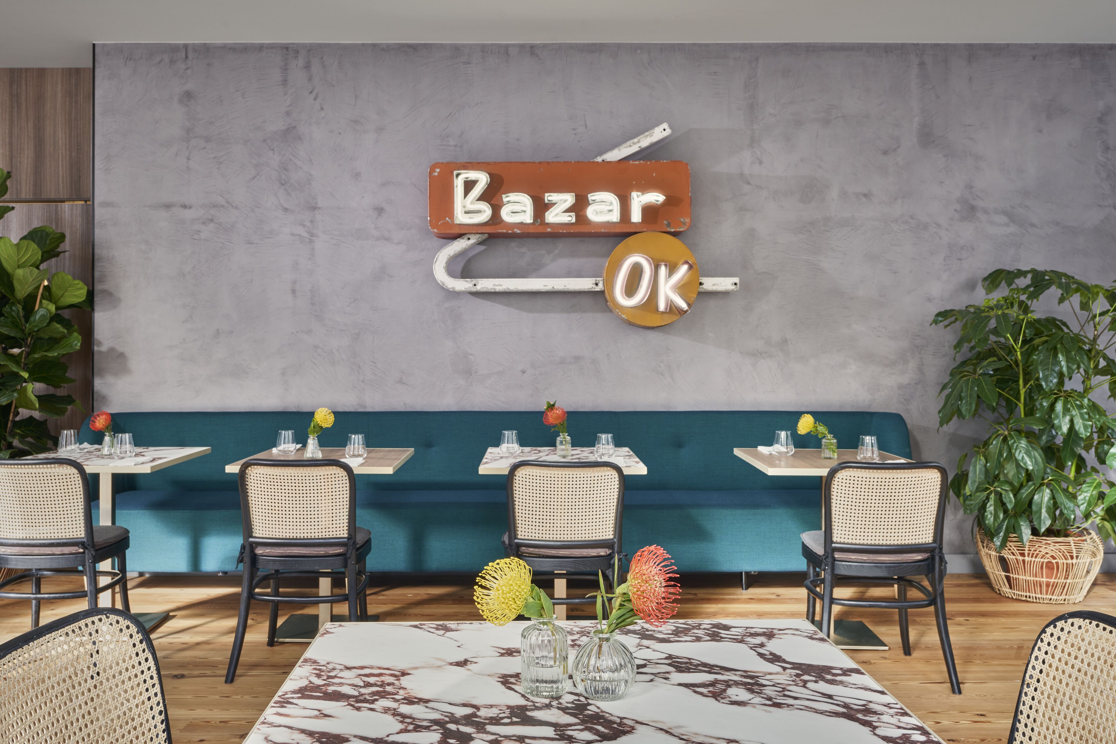 Un interior de restaurante con un letrero que dice 'Bazar OK', sillas de mimbre, mesas con manteles blancos y plantas en macetas.