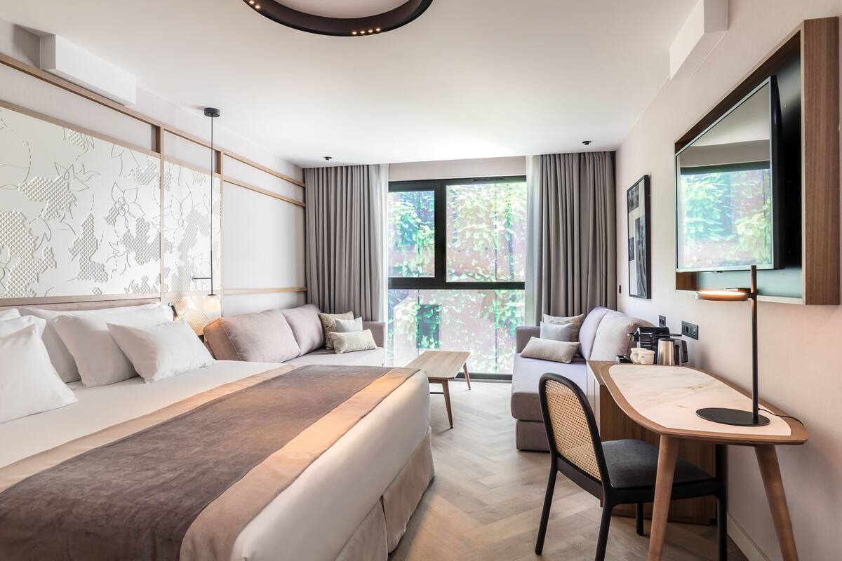 A modern hotel room with a large bed, a desk, and a view of greenery through the windows.