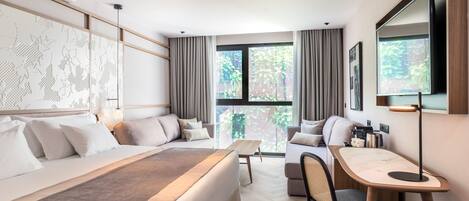 A modern hotel room with a large bed, a desk, and a view of greenery through the windows.