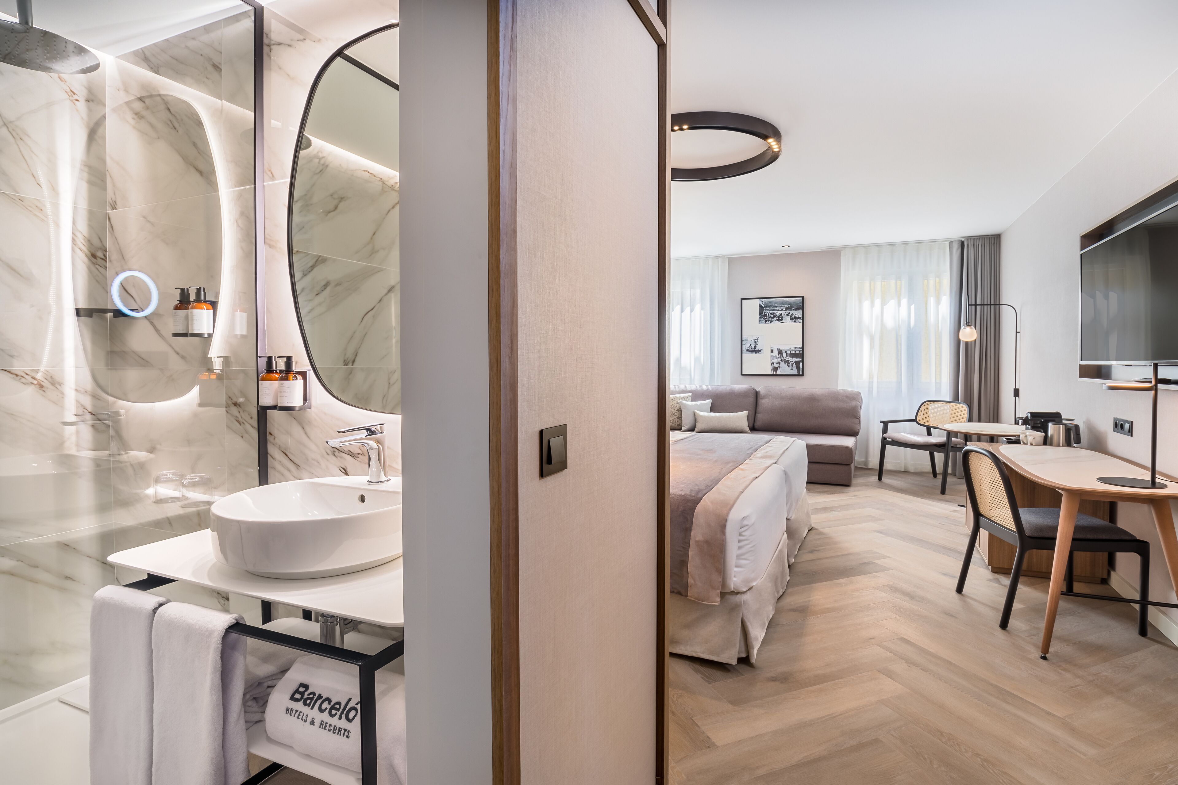 Una habitación de hotel moderna con un baño de mármol, una cama con cabecera, una zona de comedor con mesa y sillas, y un televisor.