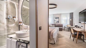 Una habitación de hotel moderna con un baño de mármol, una cama con cabecera, una zona de comedor con mesa y sillas, y un televisor.