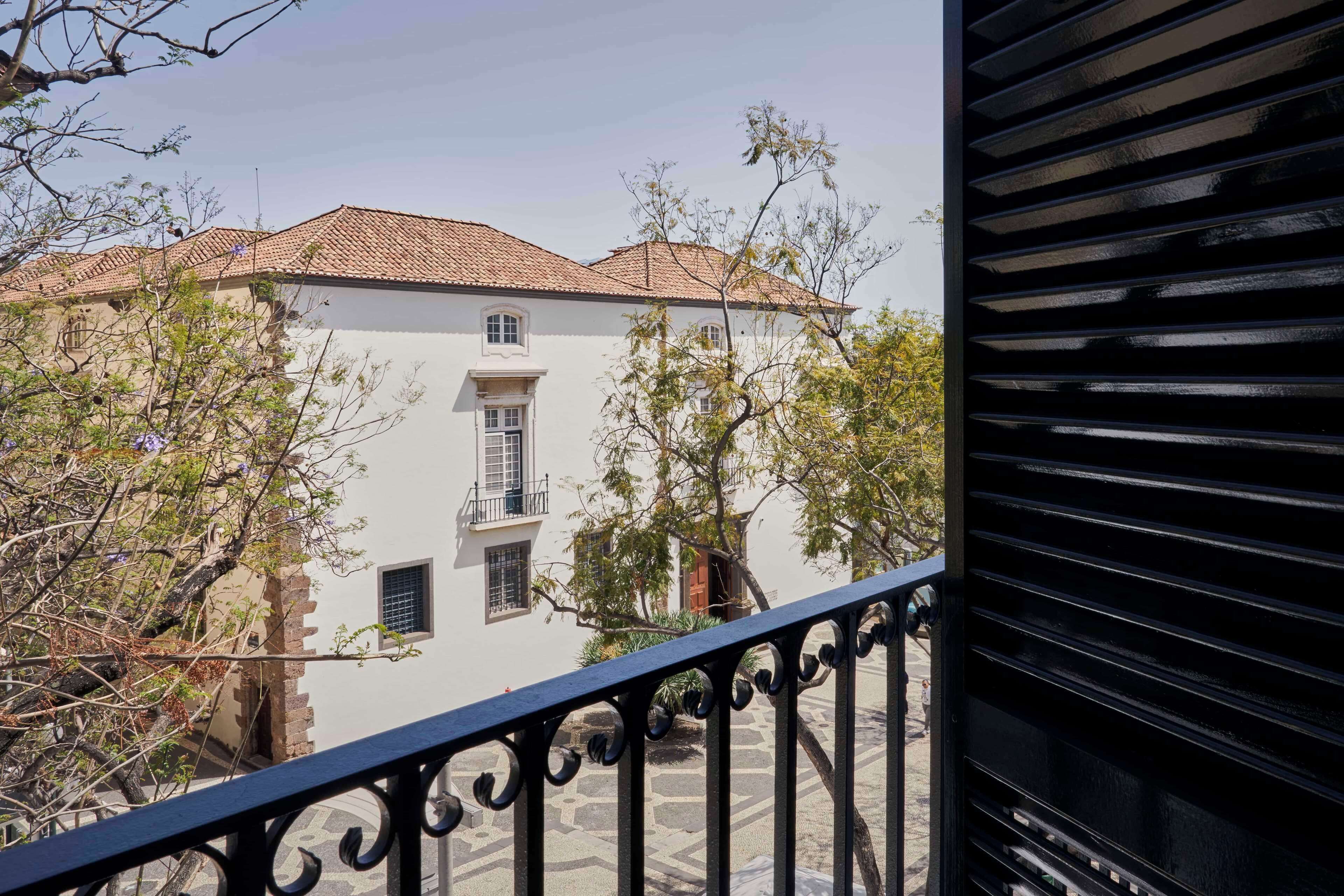 A building with a tiled roof, a balcony with ornate metal railings, and a courtyard with trees and a stone floor.