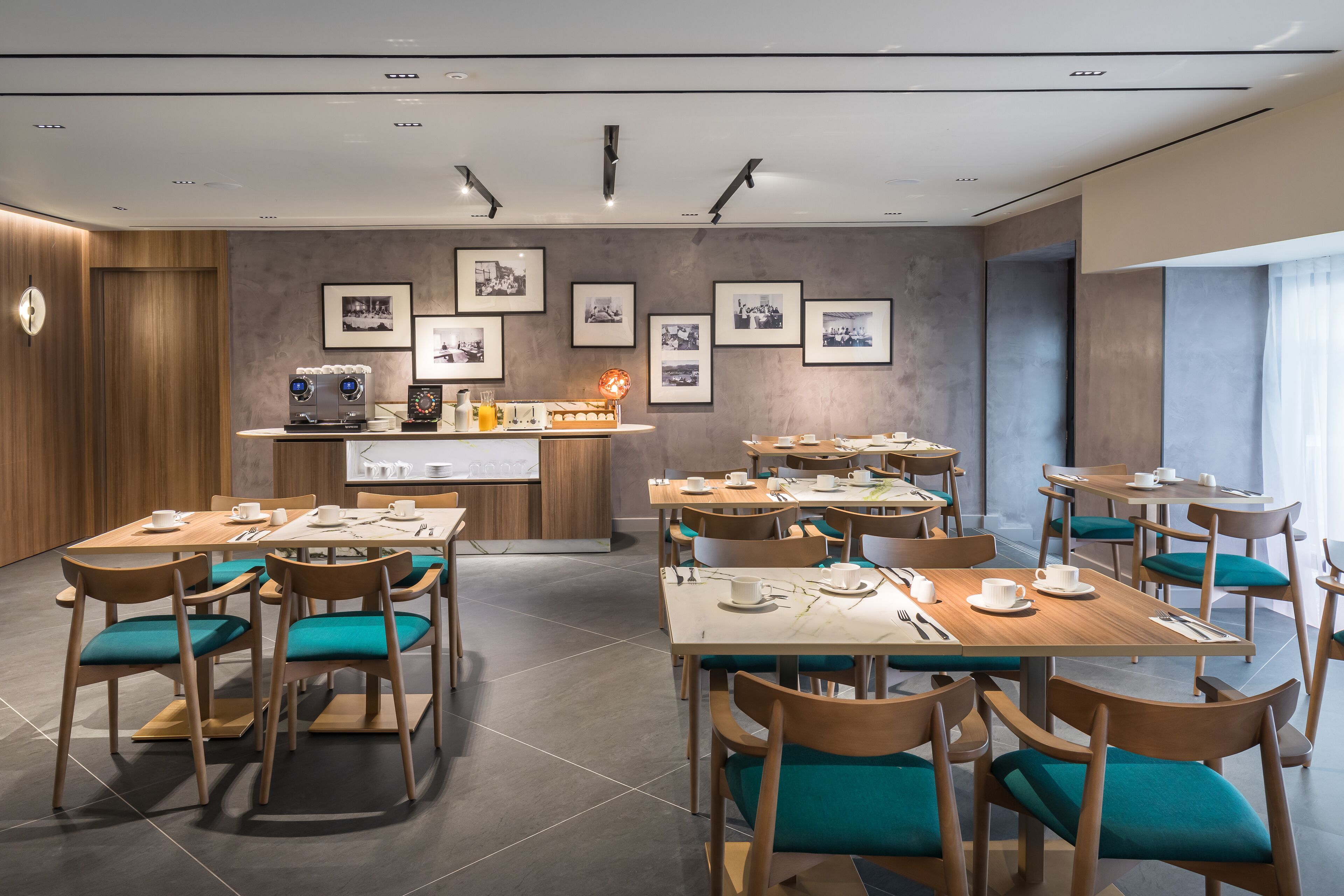 A modern dining area with wooden tables and teal chairs, a coffee station, and framed pictures on the wall.