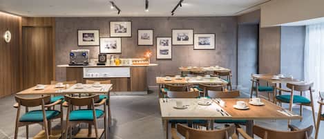 A modern dining area with wooden tables and teal chairs, a coffee station, and framed pictures on the wall.