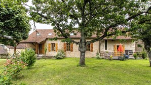 Ein Steinhaus mit einem großen Baum davor, einem grünen Rasen und einer Holzterrasse mit Gartenmöbeln.