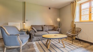 Ein Wohnzimmer mit einer Couch, zwei Sesseln, einem runden Couchtisch, einer Stehlampe und einem Fenster mit Vorhängen.