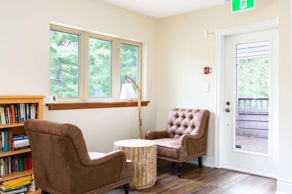 A room with a wooden bookshelf, two brown leather chairs, a round wooden table, and a window with a view of trees.