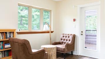 A room with a wooden bookshelf, two brown leather chairs, a round wooden table, and a window with a view of trees.