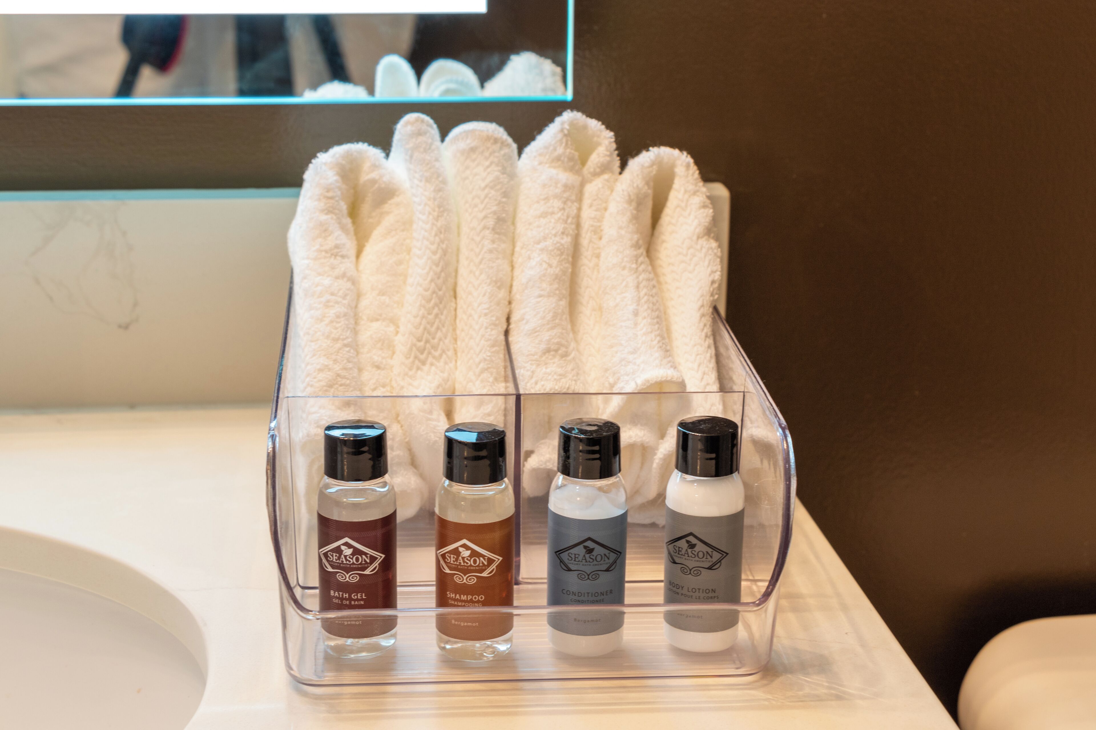 A bathroom counter with a clear plastic organizer holding four bottles labeled 'Season' with different products: bath gel, shampoo, conditioner, and body lotion.