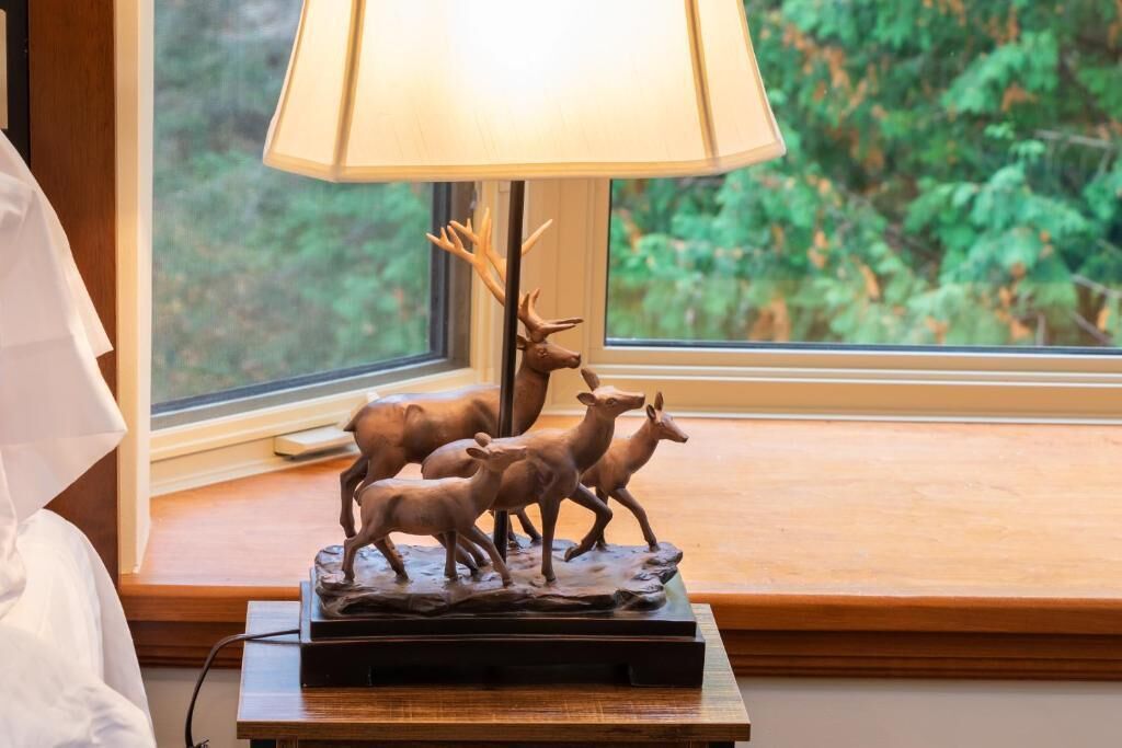 Une lampe ornée d’une figurine de cerf, posée sur une table en bois près d’une fenêtre donnant sur un paysage verdoyant.