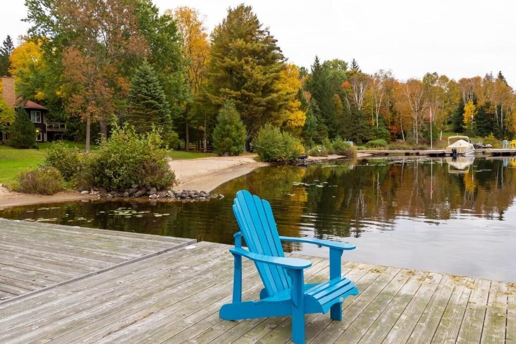 A wooden dock with a blue Adirondack chair, a calm lake, and autumn-colored trees in the background.
