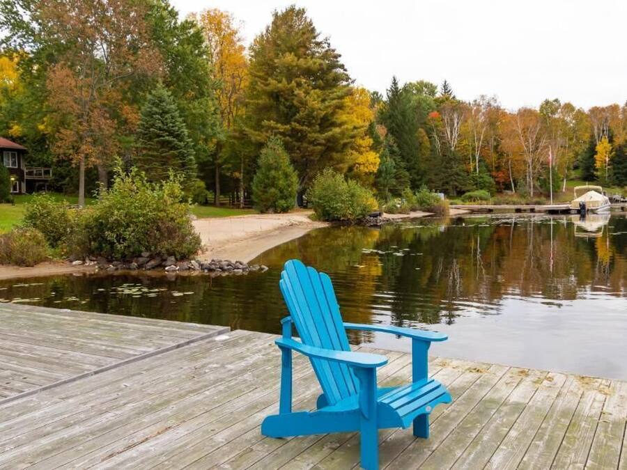 A wooden dock with a blue Adirondack chair, a calm lake, and autumn-colored trees in the background.