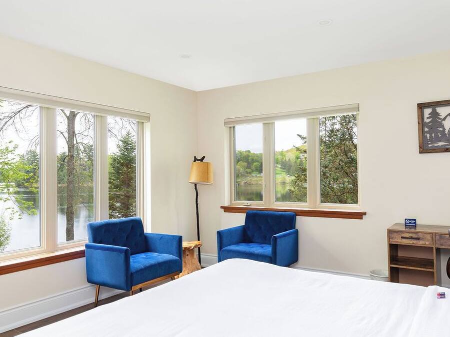 Una habitación con una cama, dos sillas azules, un escritorio y ventanas amplias con vistas a árboles y una masa de agua.