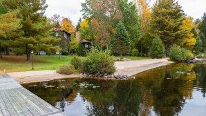 A wooden dock extending into a calm lake, surrounded by autumn-colored trees and a residential building in the background. - Fairy Bay Lakehouse (Huntsville)