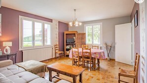 A cozy living room with a sofa, a wooden table, and a dining area with a table and chairs. There is a television on a wooden cabinet and a window with a view of greenery. - Le Champ des Charmes (SAINT-DIDIER-SUR-ARROUX)