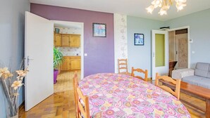 A dining area with a round table, chairs, and a colorful tablecloth. There is a kitchen visible through an open door, and a living area with a sofa and a lamp. - Le Champ des Charmes (SAINT-DIDIER-SUR-ARROUX)