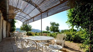 A covered outdoor seating area with a round table and chairs, surrounded by greenery and a view of hills.