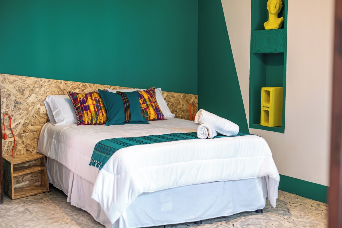 A neatly made bed with a white comforter and colorful pillows, a wooden nightstand with a lamp, and a green accent wall with a yellow shelf.