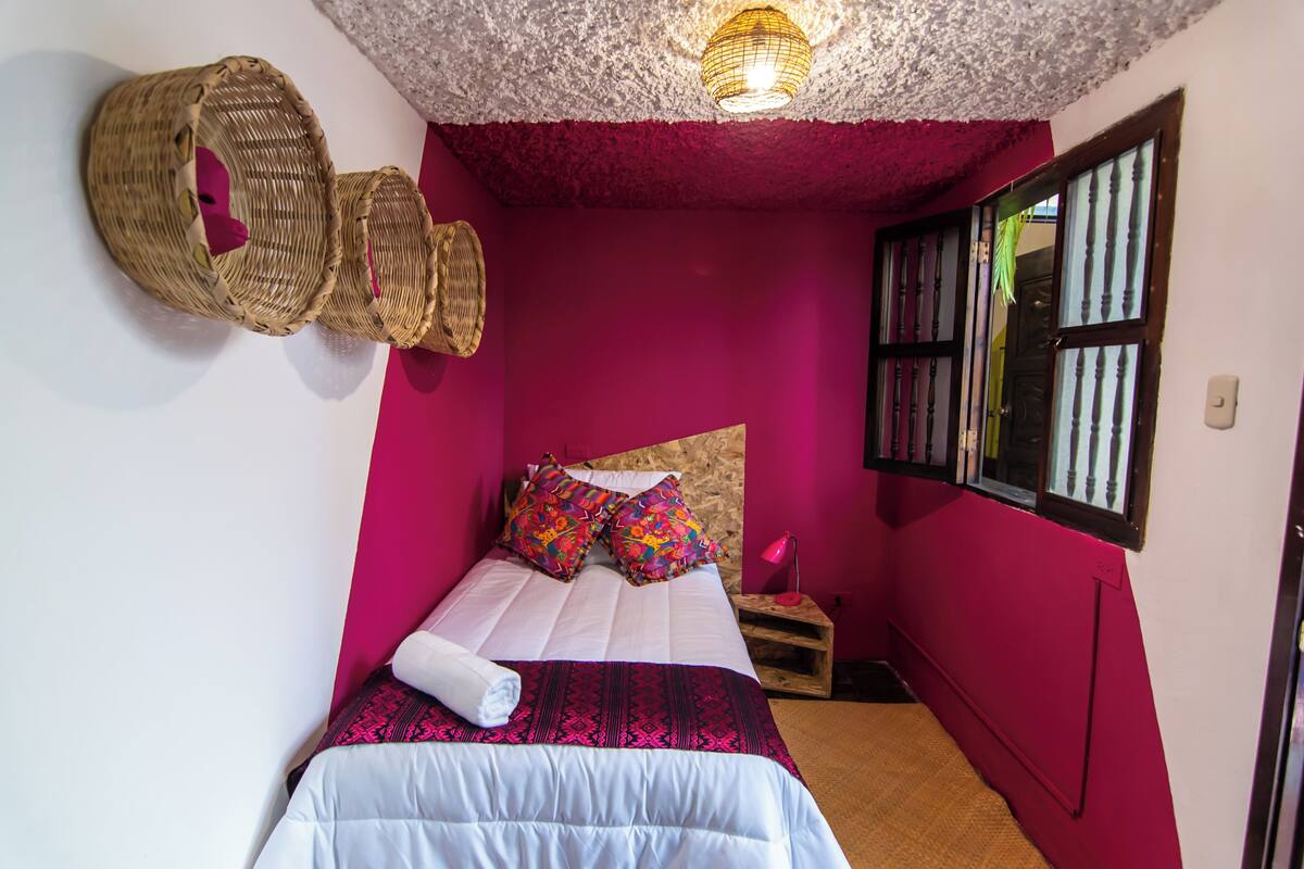 A room with a bed, a nightstand, a wicker basket, a window with blinds, and a textured ceiling.
